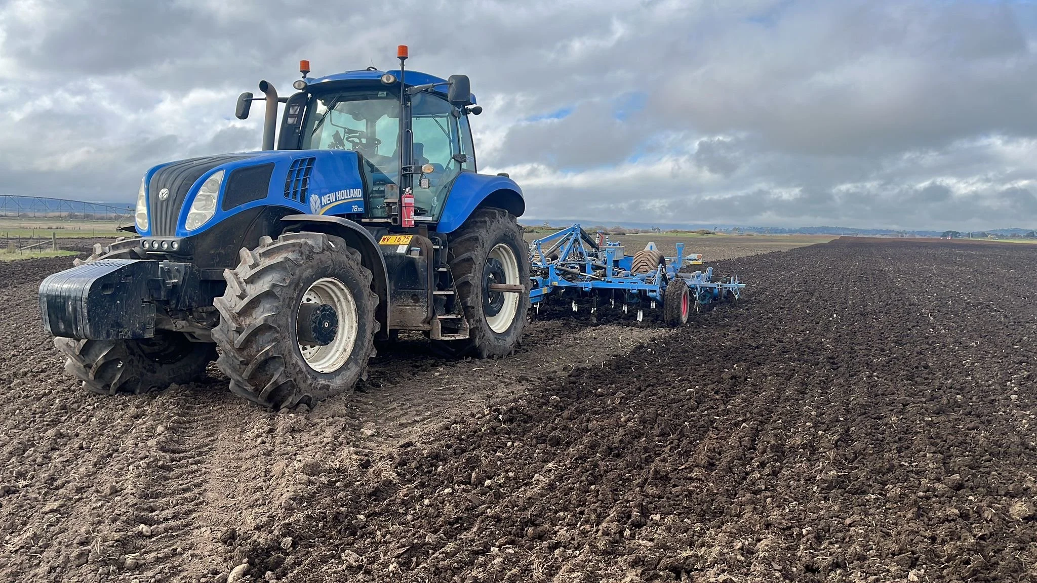 A blue New Holland tractor plowing a large field of dark soil, with an overcast sky and distant horizon.
