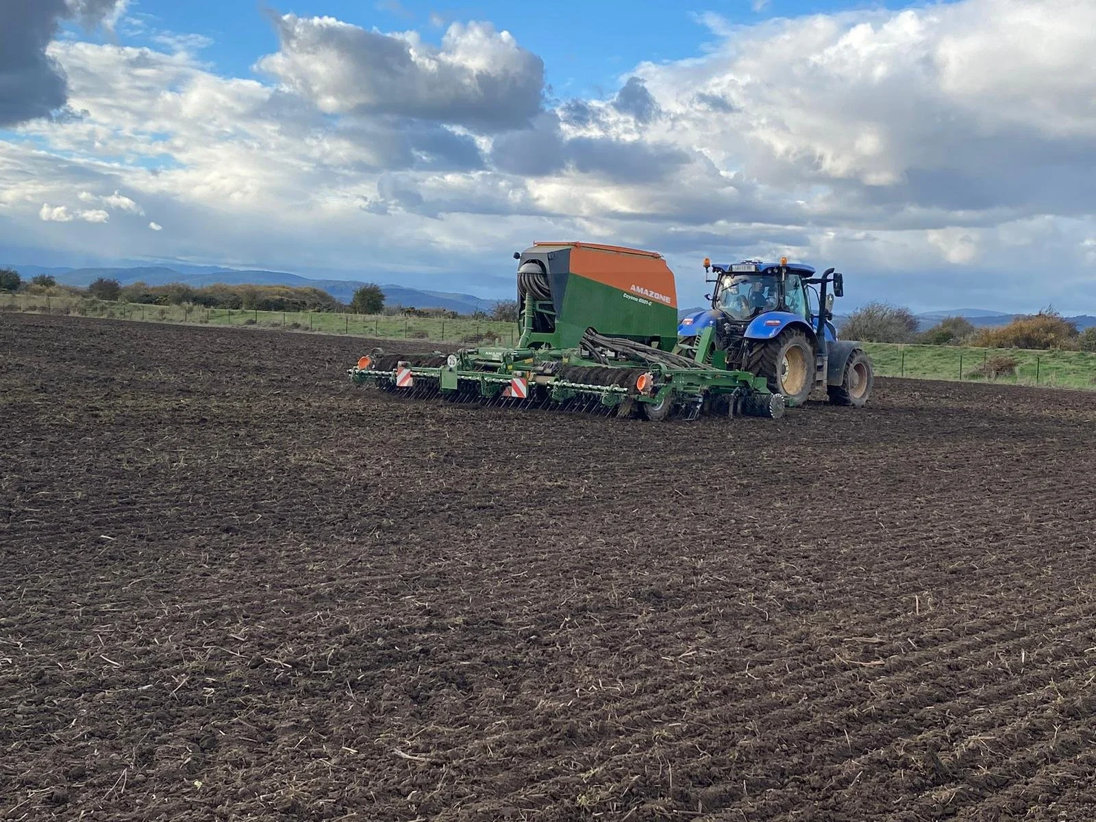 A tractor with a planting or tilling attachment working in a large, open farm field under a cloudy sky.