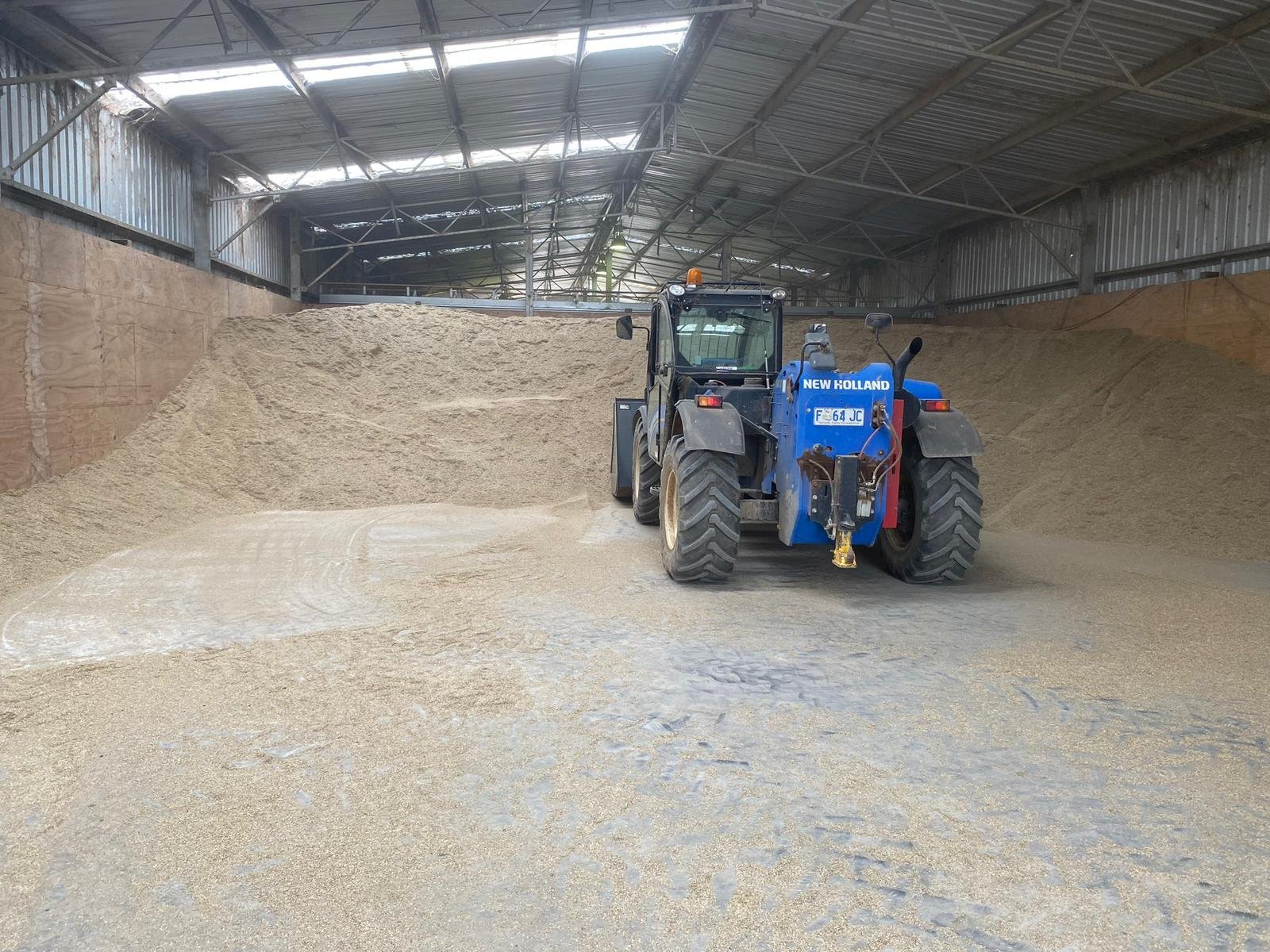 Inside a large warehouse or storage area with a blue New Holland tractor moving or spreading a large amount of grain or sand, surrounded by walls made of metal and wood, with a high, partially translucent roof letting in natural light.