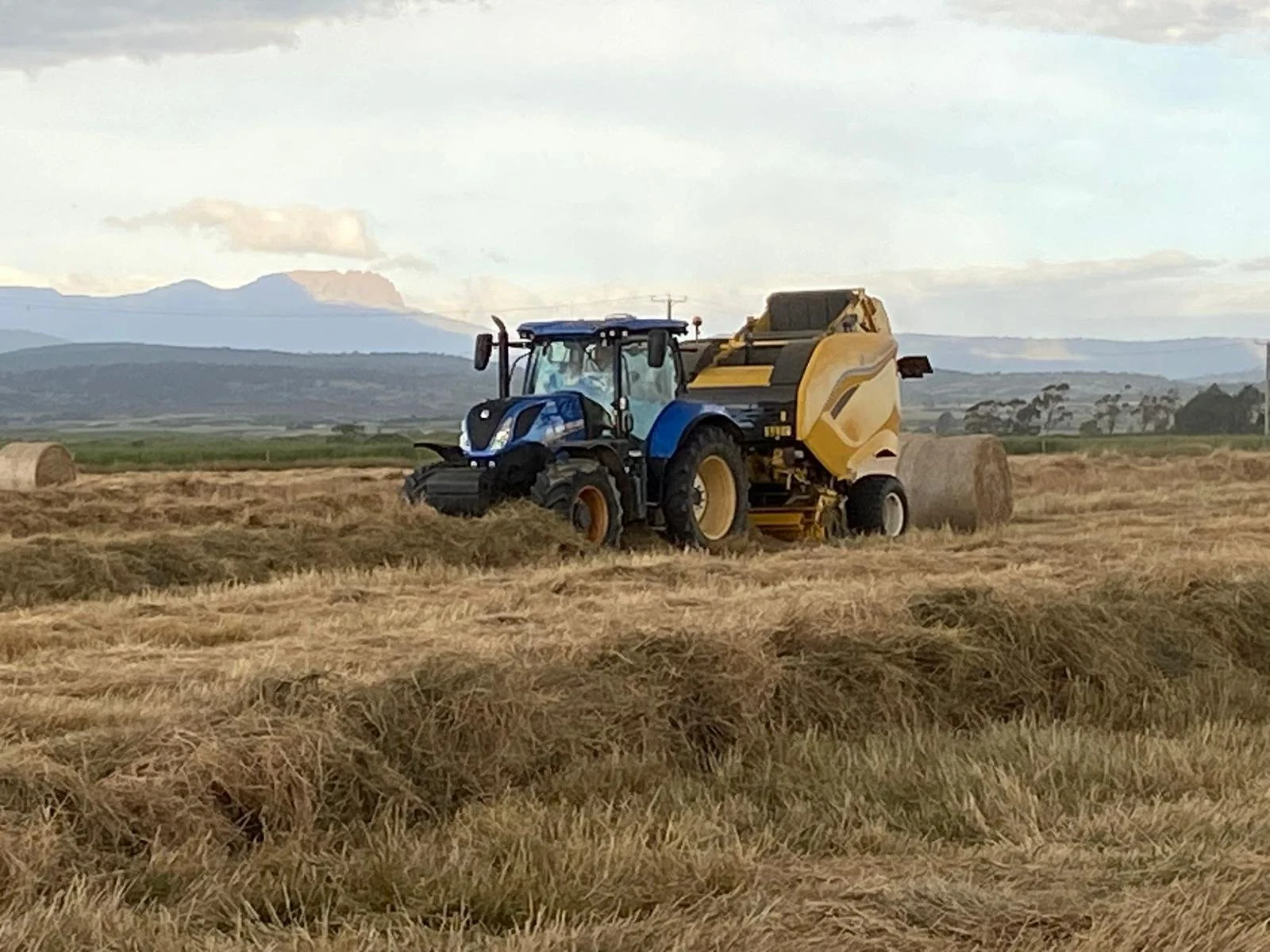 A blue tractor with a yellow baler attached is harvesting hay in a field with hay bales scattered across the landscape. Mountains are visible in the background under a partly cloudy sky.