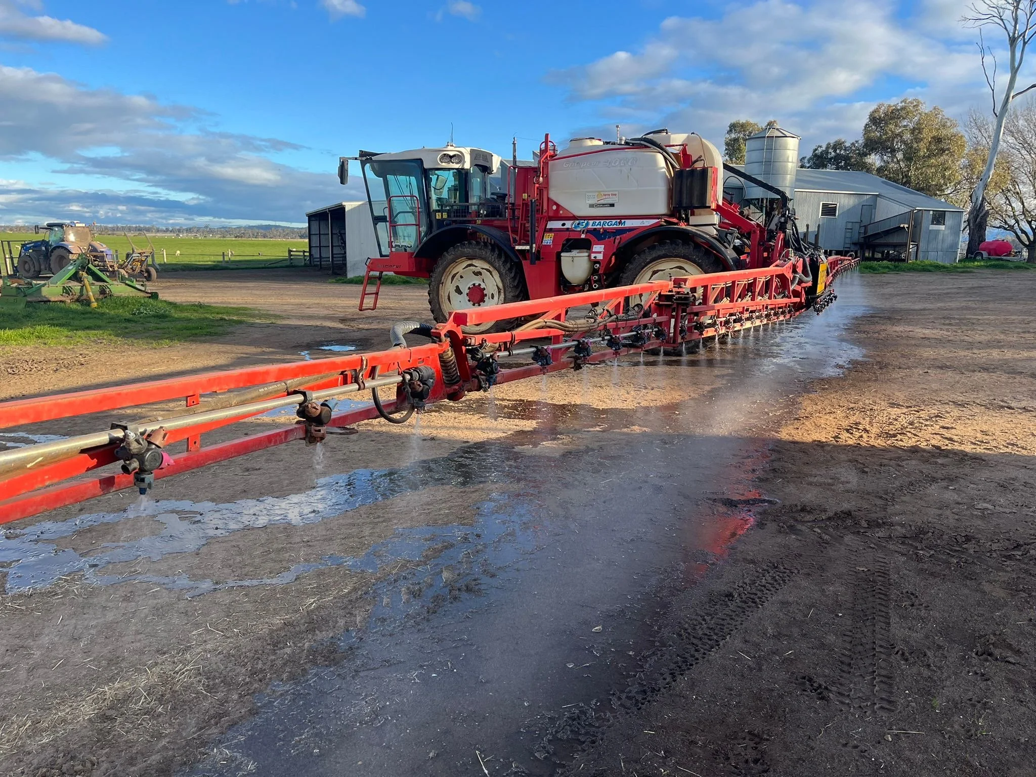 A red tractor spraying liquid on a farm field, with a barn, other tractors, and farm equipment in the background.