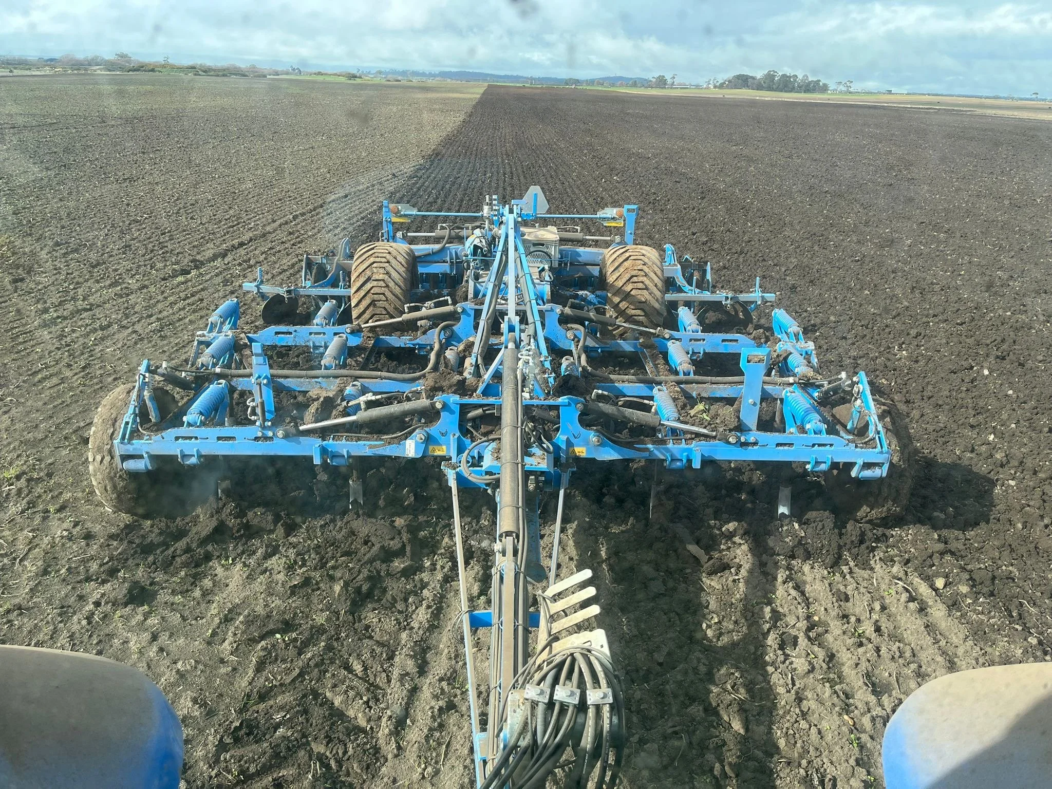 A blue tractor attachment cultivating a large field of dark, freshly tilled soil on a partly cloudy day.