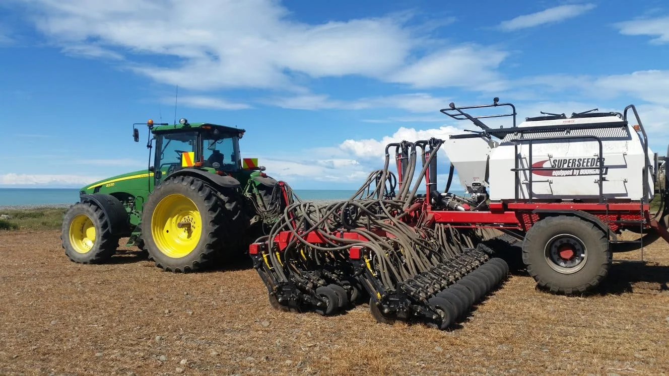 A green tractor pulling a large white and red agricultural sprayer with multiple hoses on a dirt field, blue sky, and ocean in the background.