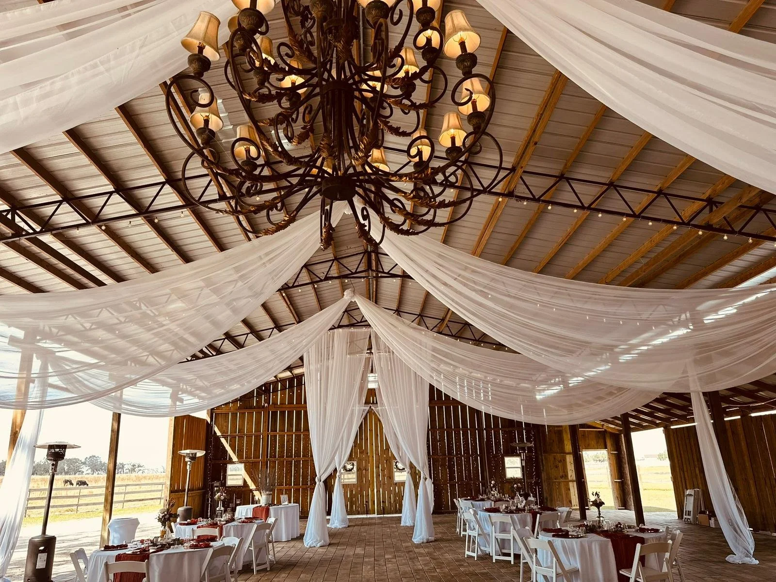 Interior of a rustic barn decorated for a wedding or event, with white draped fabric hanging from the ceiling and a large black chandelier.