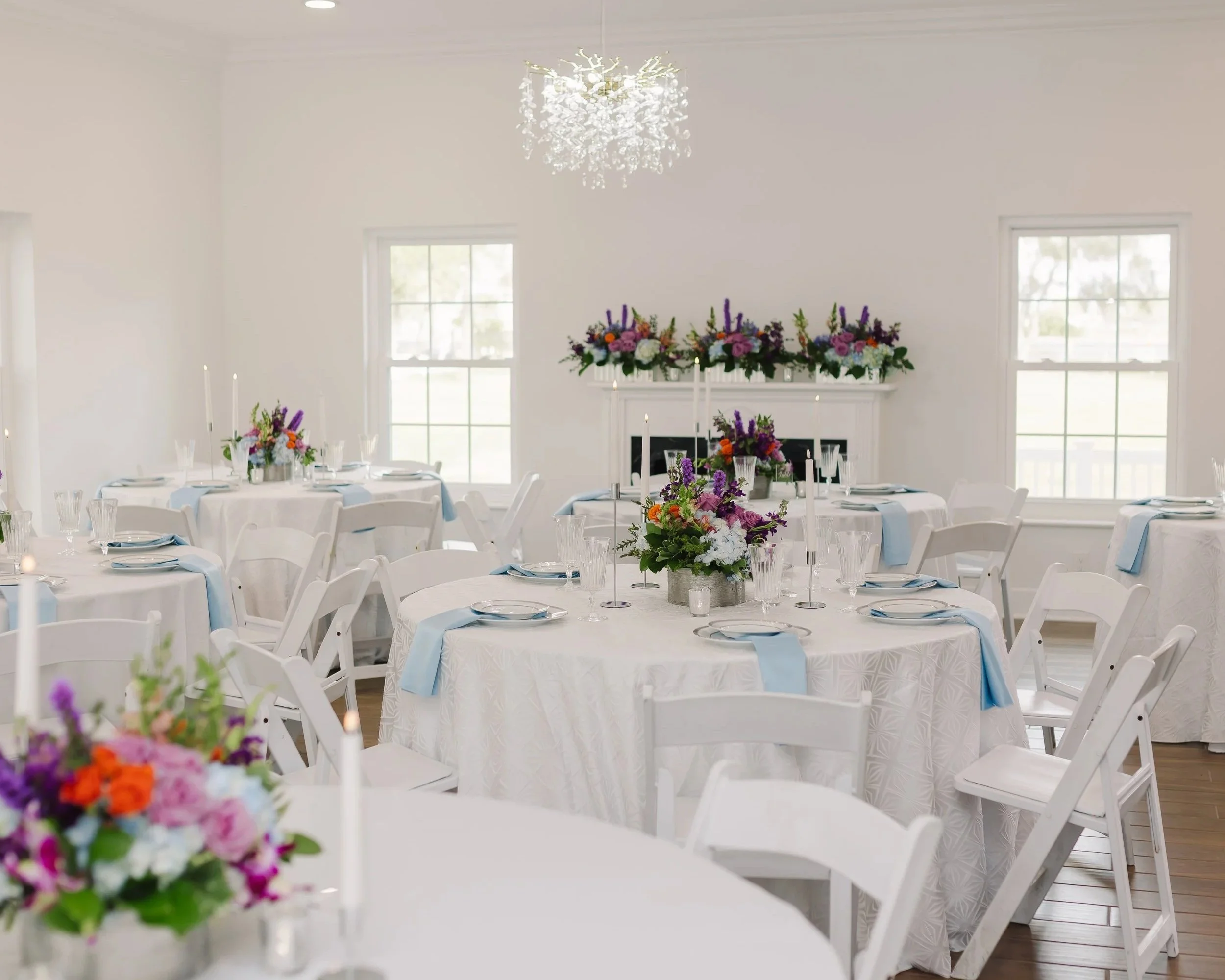 Elegant event space with round tables covered in white tablecloths, decorated with floral centerpieces and tall candles, set for a formal occasion in a bright room with large windows and a chandelier.