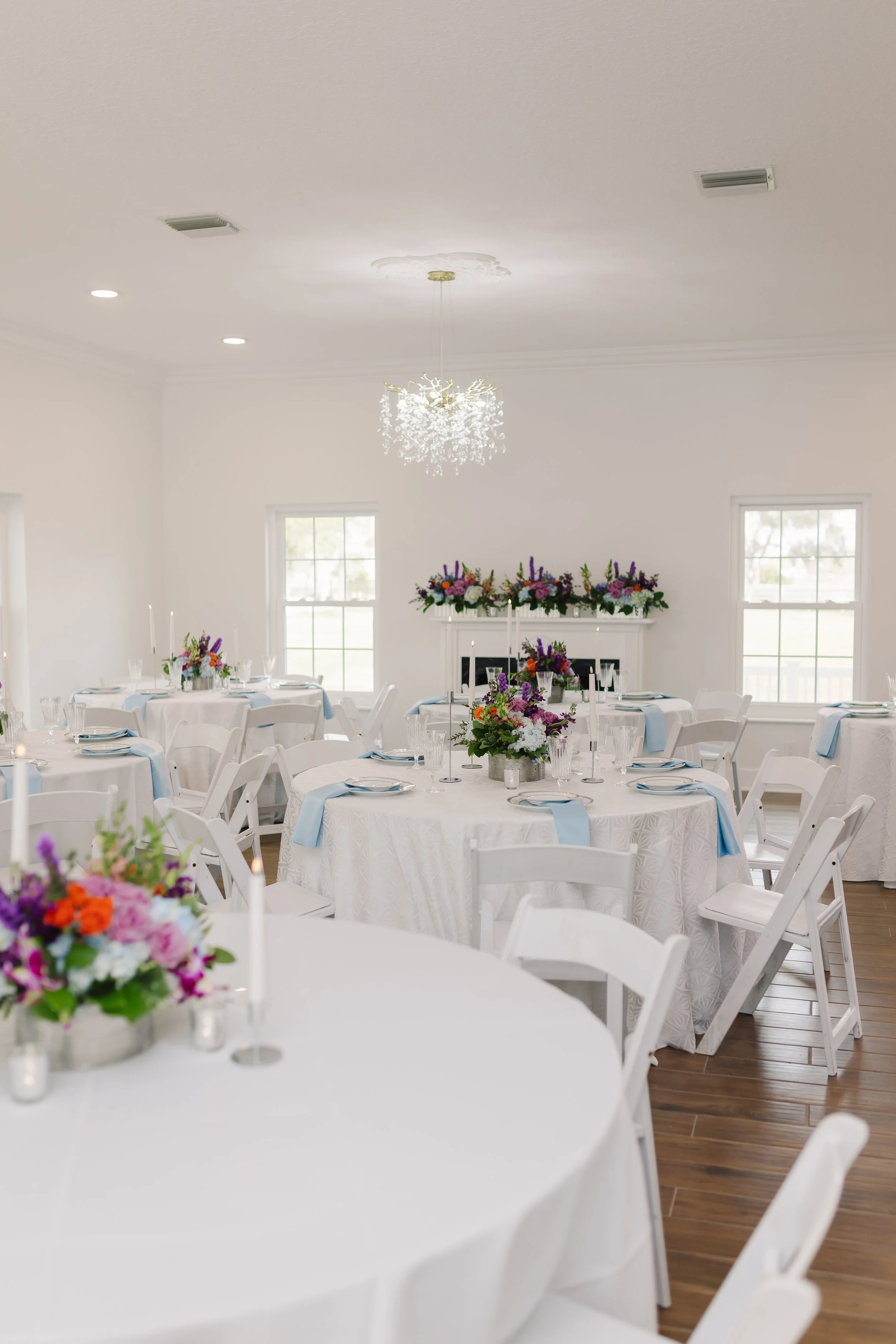 Elegant event reception decorated with white tablecloths, colorful floral centerpieces, and light blue napkins, arranged in a bright room with large windows and a chandelier.
