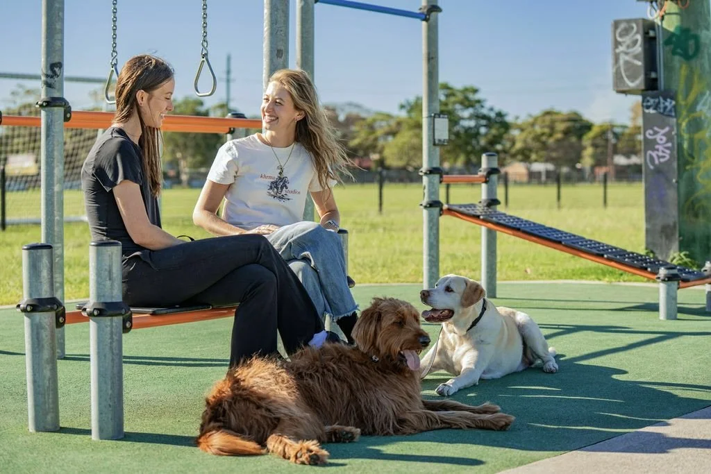 Two women sitting on a park bench at a playground, smiling and talking, with two dogs lying on the ground nearby, in an outdoor park setting.