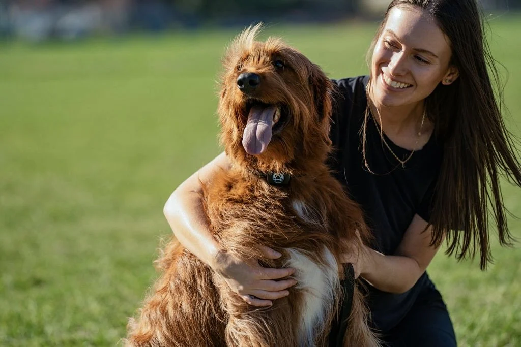 A woman smiling and hugging her large, fluffy brown dog with its tongue out, on a grassy field.