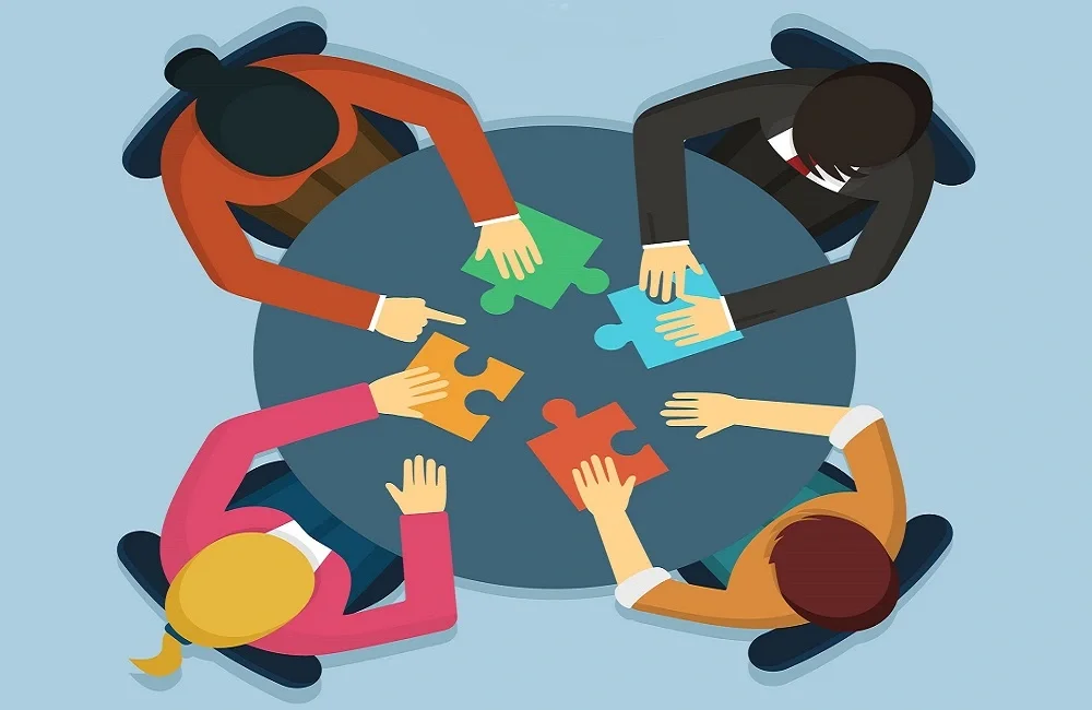 Quatro pessoas com diferentes roupas montando um quebra-cabeça na mesa. Cada pessoa segura uma peça de uma cor diferente, simbolizando colaboração.