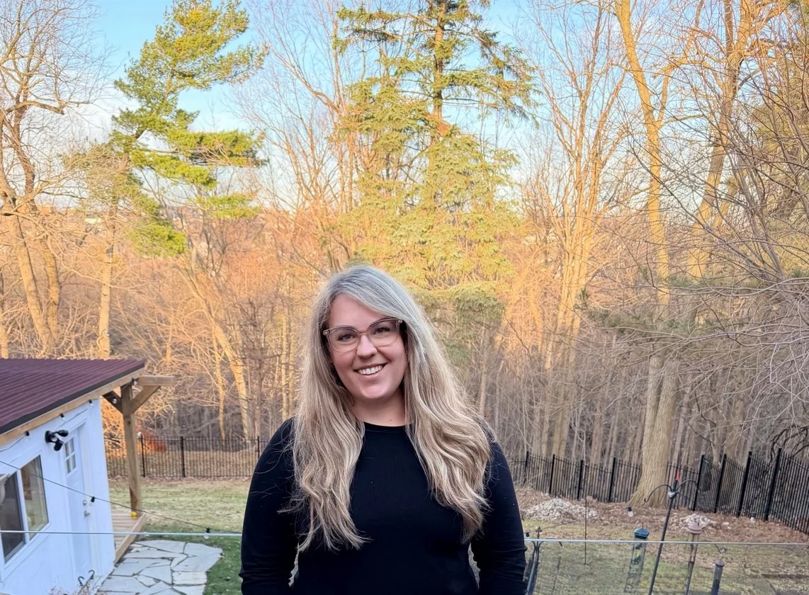 Kassandra Smalley, a woman with long blonde hair, glasses, and a black top smiling outdoors in a backyard with trees and a fence.