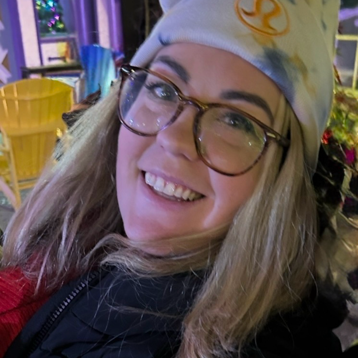 Kassandra Smalley, a close-up of a smiling woman wearing glasses with a beanie hat, outdoors at night with colorful lights and outdoor furniture in the background.
