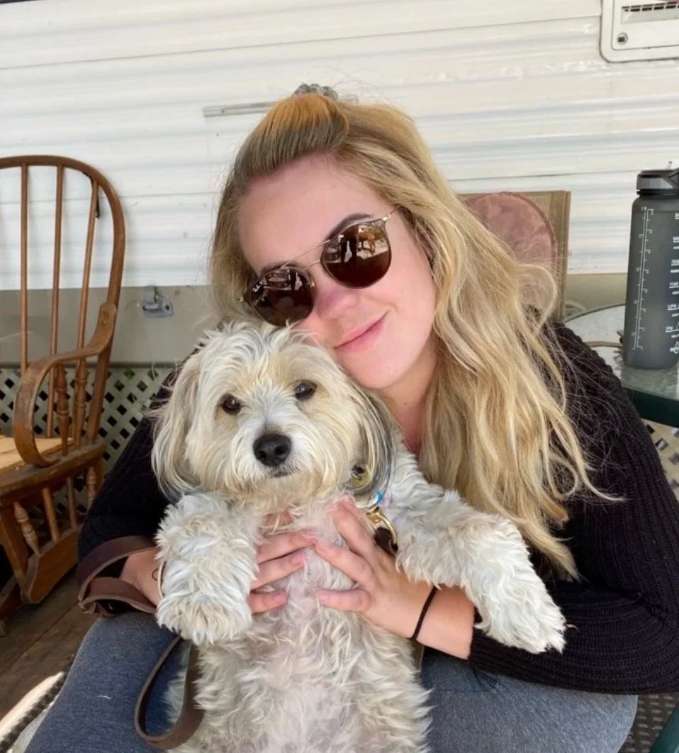Kassandra Smalley, a woman with long blonde hair, wearing sunglasses, smiling and holding a fluffy white dog in her arms. They are sitting outside on a porch.