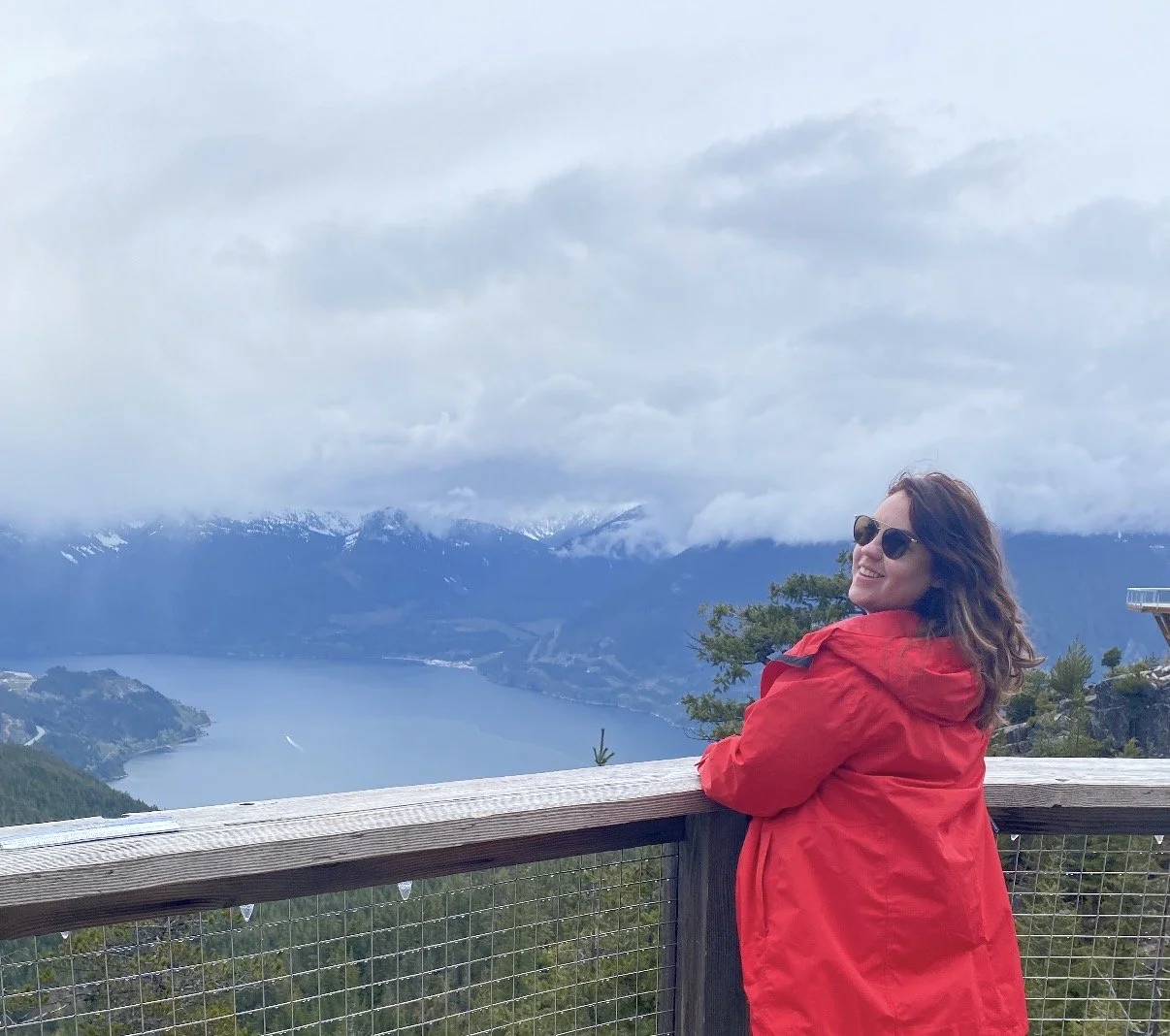 Kassandra Smalley, a young woman wearing red jacket and sunglasses smiling while leaning on a wooden railing at a scenic overlook with a lake, mountains, snow-capped peaks, and cloudy sky in the background.