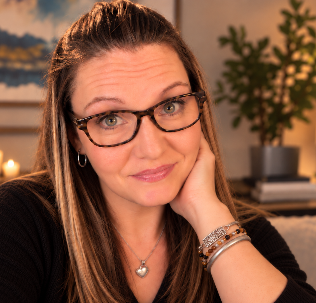 A woman with glasses and long hair, smiling softly while resting her hand on her cheek, in a cozy indoor setting with a plant and candle in the background.