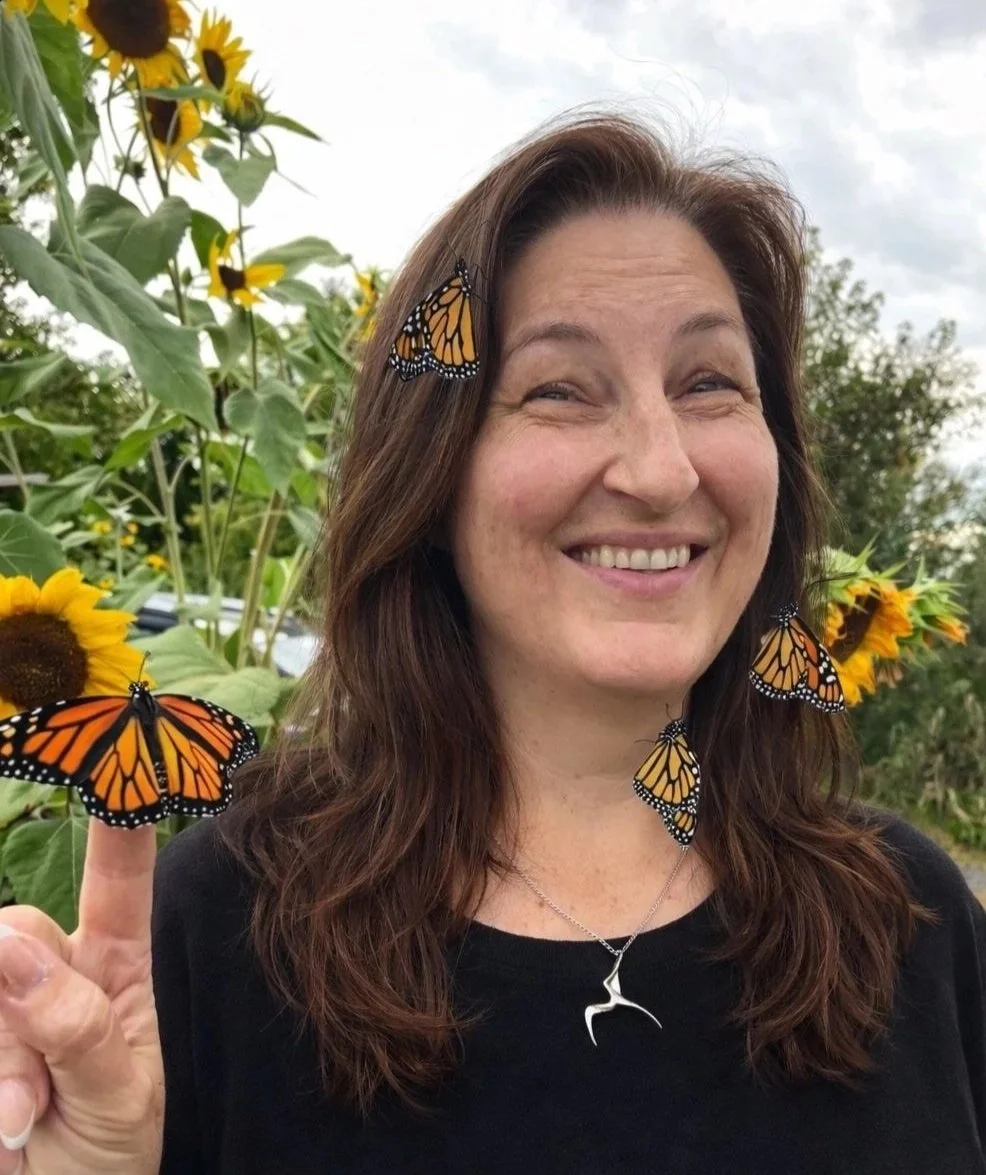 Nicola Wolters- Registered Psychotherapist (Qualifying) smiling outdoors with sunflowers in the background, having butterflies on her face and hand, including a Monarch butterfly on her finger.