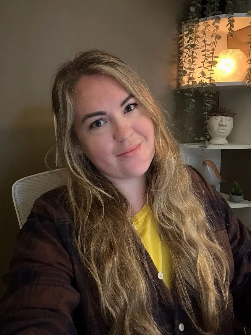Kassandra Smalley, a woman with long wavy blonde hair, wearing a brown plaid shirt over a yellow top, sitting in a room with soft lighting, a decorative shelf with plants, a lamp, and a sculpture of a face.