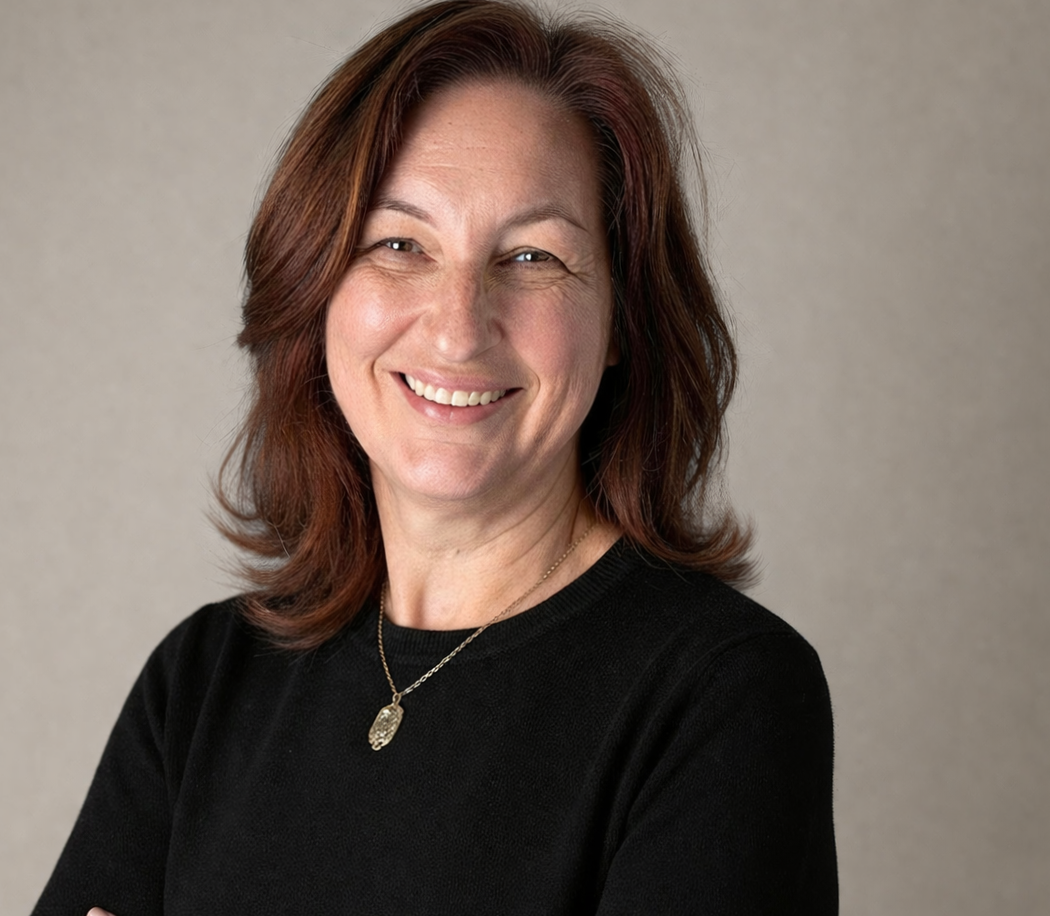 Close-up of Nicola Wolters- Registered Psychotherapist (Qualifying) with shoulder-length reddish-brown hair, wearing a black top and a gold necklace, against a neutral background.