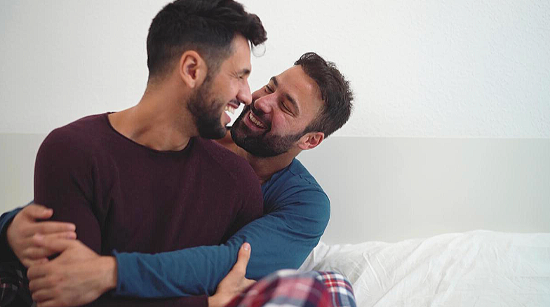 Two men sitting on a bed, hugging, and smiling at each other.