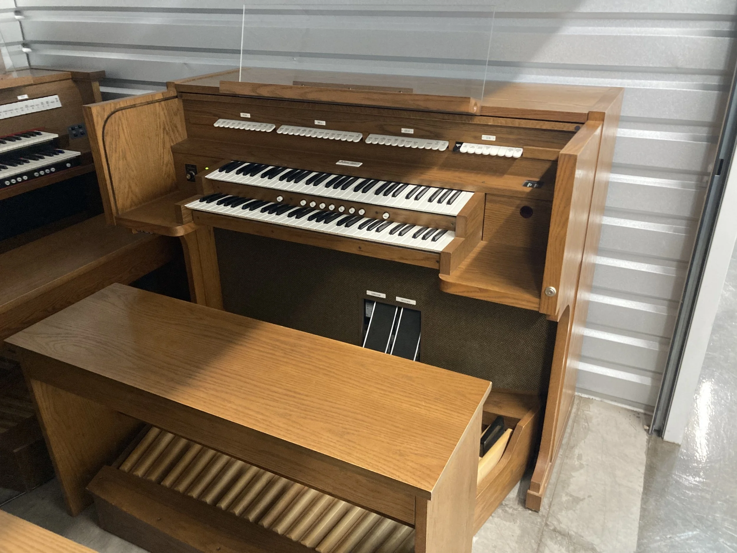 a picture of a mahogany colored digital church organ with two manual keyboards, a pedalboard, two swell shade pedals, and numerous white buttons for the variety of stops it can play. The organ is positioned in front of a white metal roll-top door.