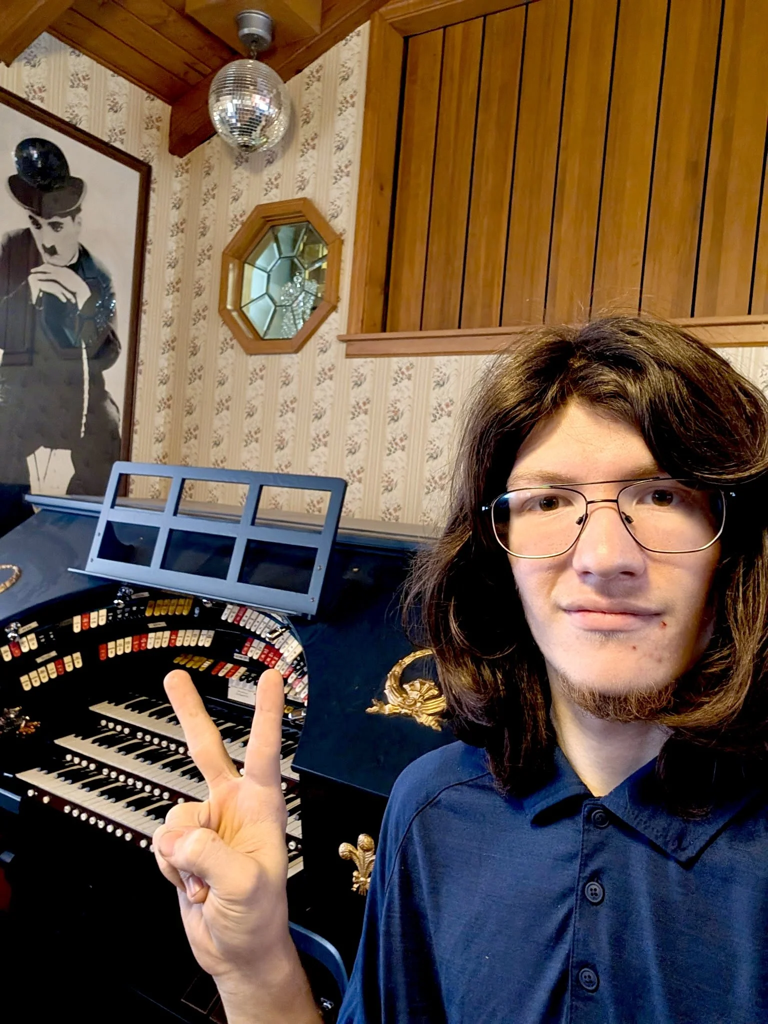A young man with glasses and shoulder-length brown hair is taking a selfie in a room decorated with vintage wallpaper and wooden paneling. He is making a peace sign with his right hand. Behind him is a black and white portrait of Charlie Chaplin, a small octagonal mirror, and a disco ball hanging from the ceiling. There is also a display of various small objects and a keyboard instrument.