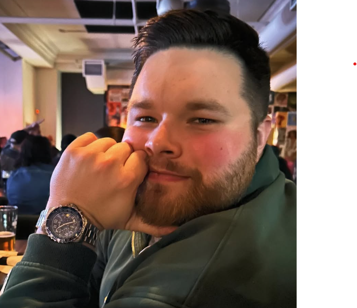 A young man with a beard and short dark hair, smiling and resting his head on his hand in a restaurant or bar. He is wearing a dark green jacket and a silver watch.