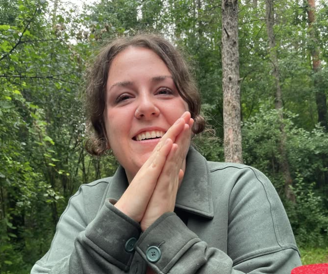 A young woman with short curly brown hair smiling and holding her hands together near her cheek outdoors in a forested area with green trees.