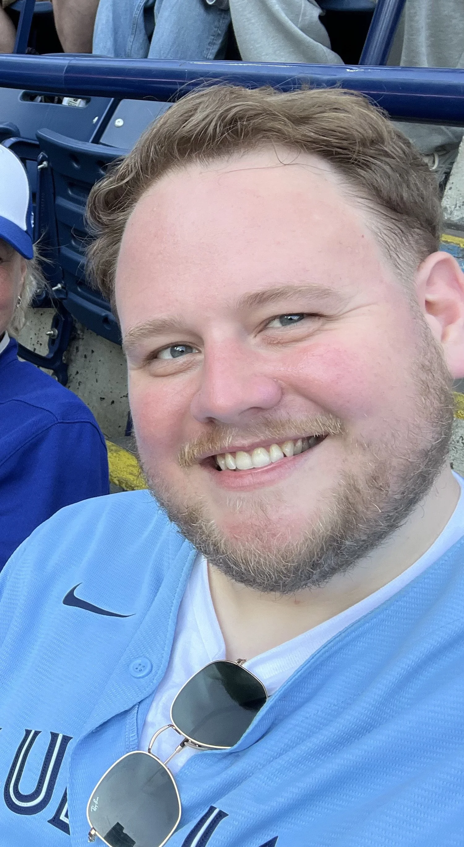 A smiling man with light brown hair and a beard wearing a light blue sports jersey and sunglasses hanging from his collar, sitting in a stadium.
