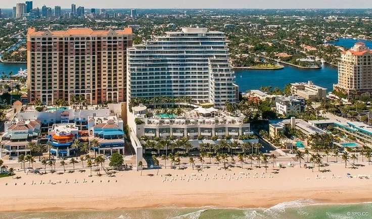 Aerial view of a beachside city with high-rise buildings, hotels, and condominiums along the shoreline, with water bodies and boats in the background.