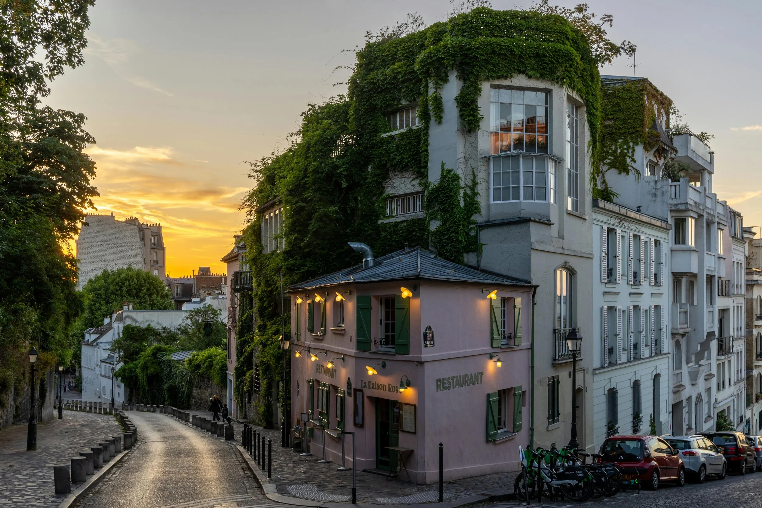 Rue pavée avec un bâtiment de restaurant au coin, entouré d'arbres et de bâtiments résidentiels, au coucher du soleil.
