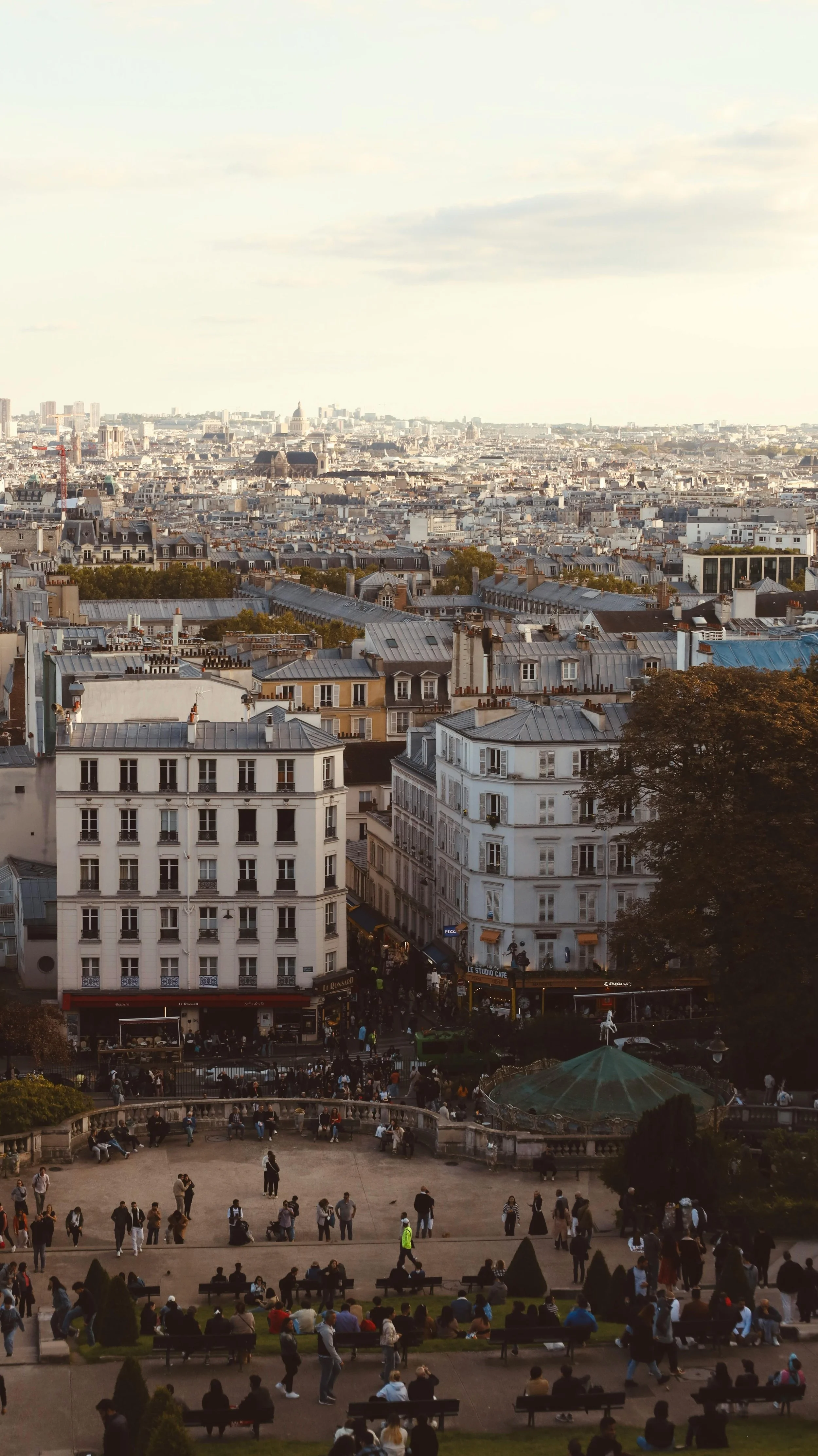 Vue d'une ville européenne avec des bâtiments résidentiels, un parc public avec des gens assis et une grande foule au bas de l'image, des arbres et un ciel nuageux.