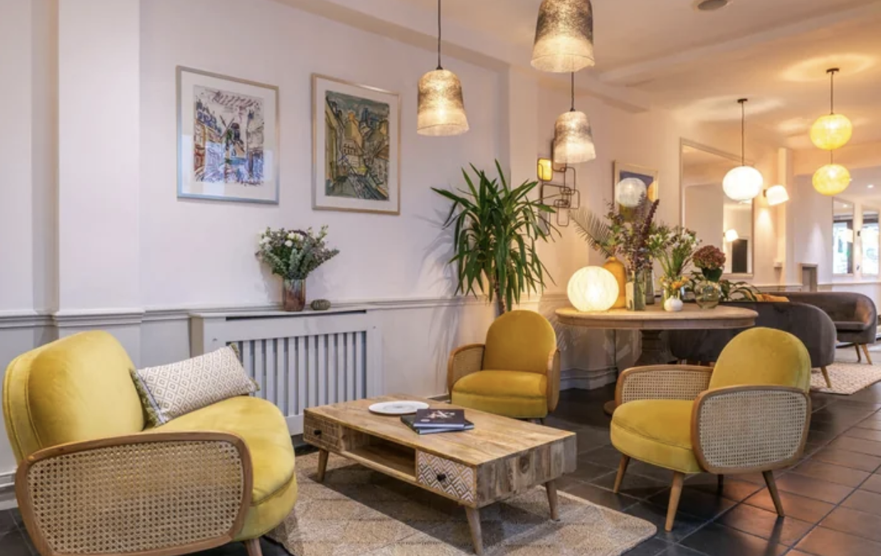 Salon avec fauteuils jaunes, table en bois, lampes suspendues, décor floral et œuvres d'art sur le mur.