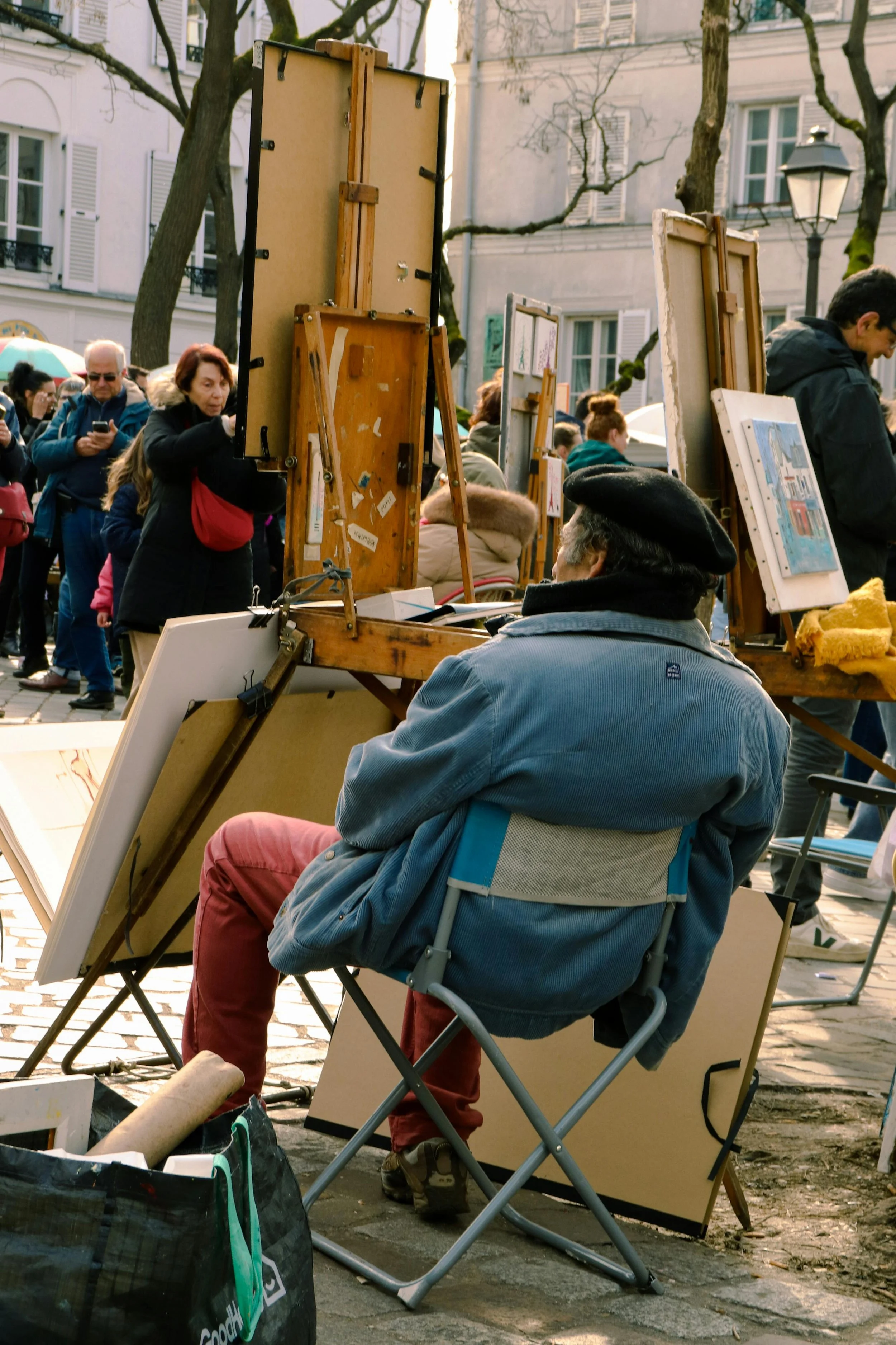 Un homme assis en plein air, probablement lors d'un marché ou d'une foire, entouré d'œuvres d'art exposées sur des chevalets, avec plusieurs personnes en arrière-plan