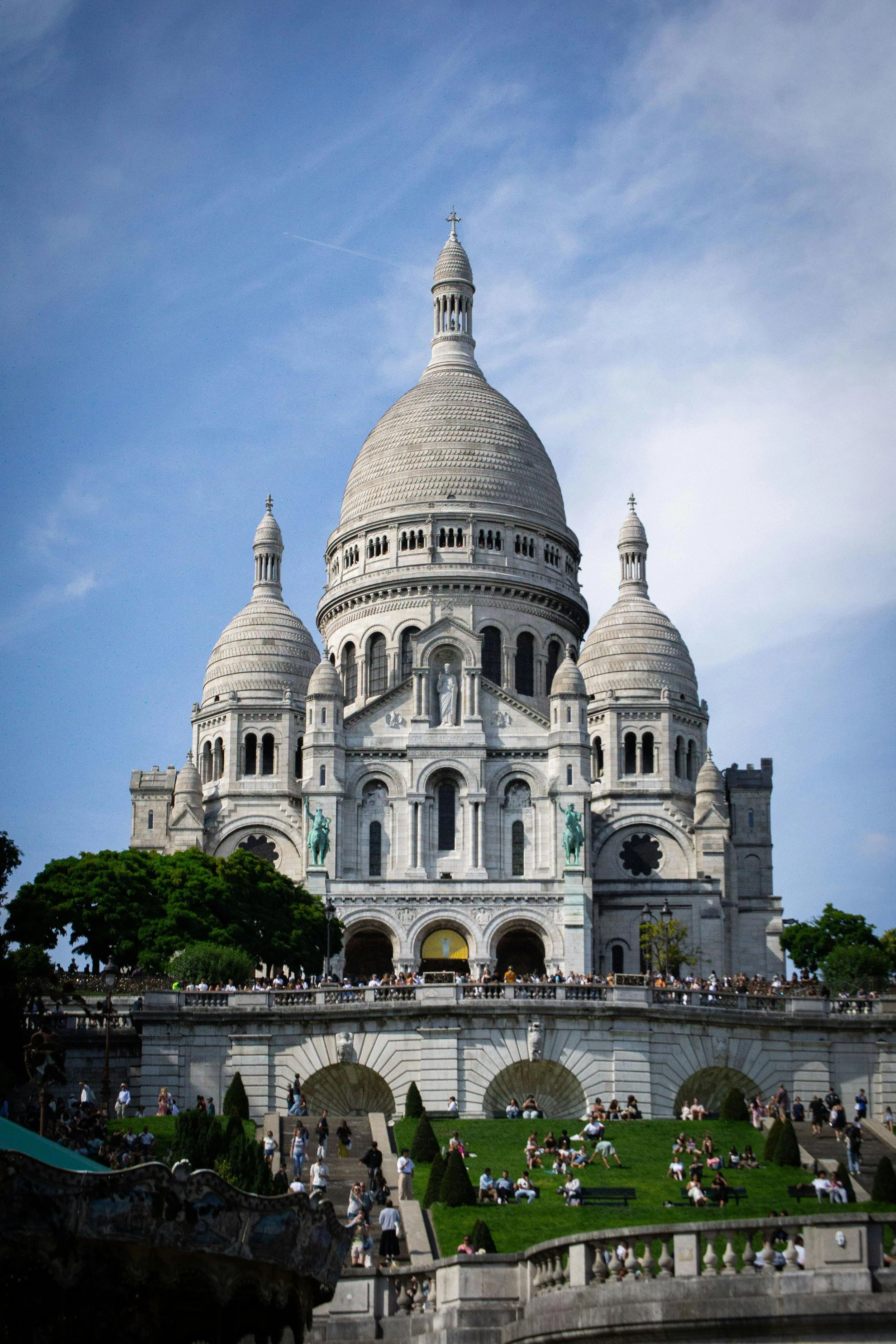 La basilique du Sacré-Cœur à Paris vue de face, avec des touristes et des visiteurs dans le parc devant, sous un ciel bleu avec quelques nuages.
