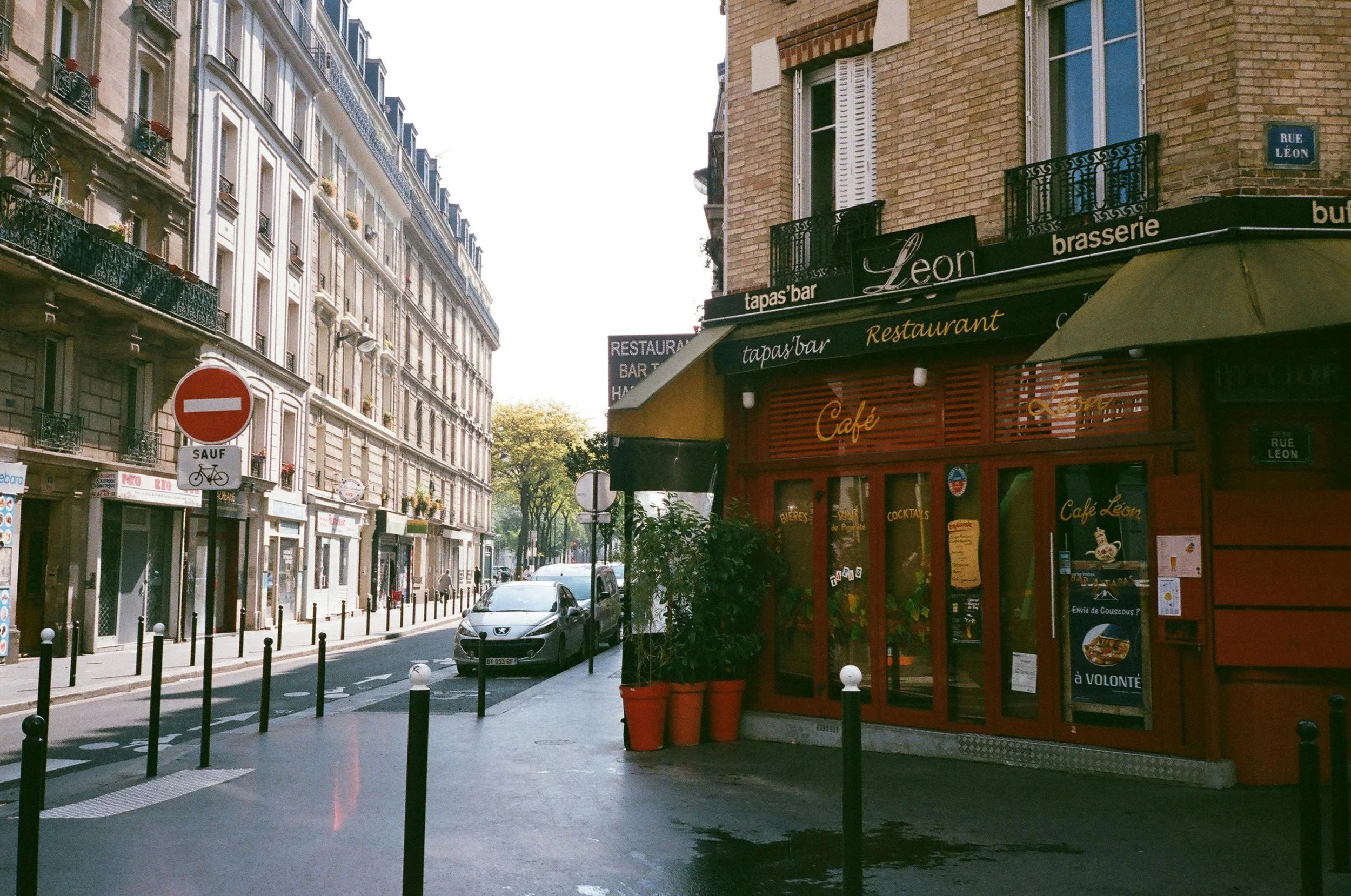 Rue de Paris avec un restaurant appelé 'León', voitures stationnées, panneaux de signalisation, immeubles résidentiels avec balcons, arbres en arrière-plan.