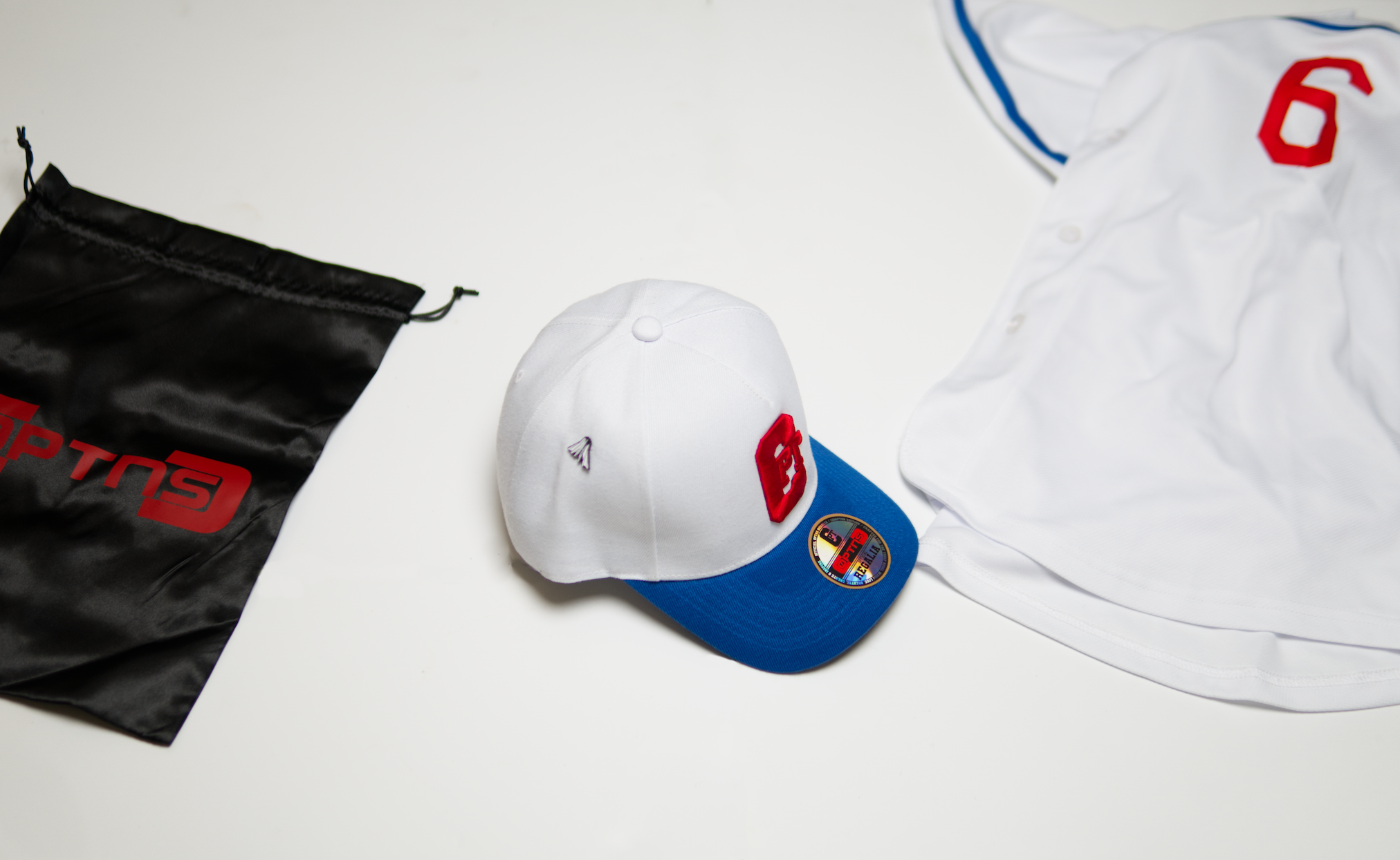 A white baseball cap with a red logo and a colorful sticker, a black bag with red logo, and a white sports jersey with red and blue accents on a white surface.