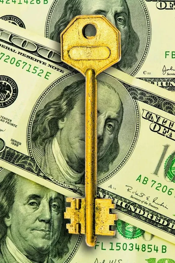 A gold-colored key placed on top of multiple 100-dollar bills featuring Benjamin Franklin's portrait.