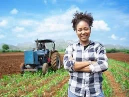 Young woman smiling with arms crossed on a farm, with a tractor and cultivated field in the background.