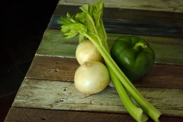 Green bell pepper, two onions, and celery stalks on a wooden surface.