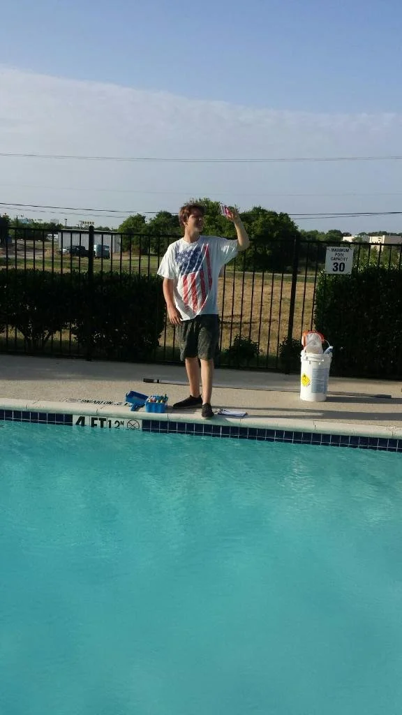A young boy stands poolside, testing pool chemicals, with a fenced outdoor area, green trees, and a cloudy sky in the background.