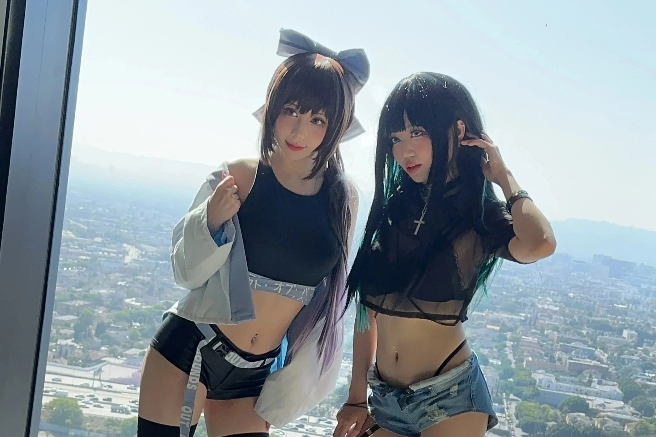 Two young women with dark hair posing in front of a tall glass window with a cityscape view in the background. They are wearing revealing clothing and accessories, with one sporting a bow headband and the other a cross necklace.