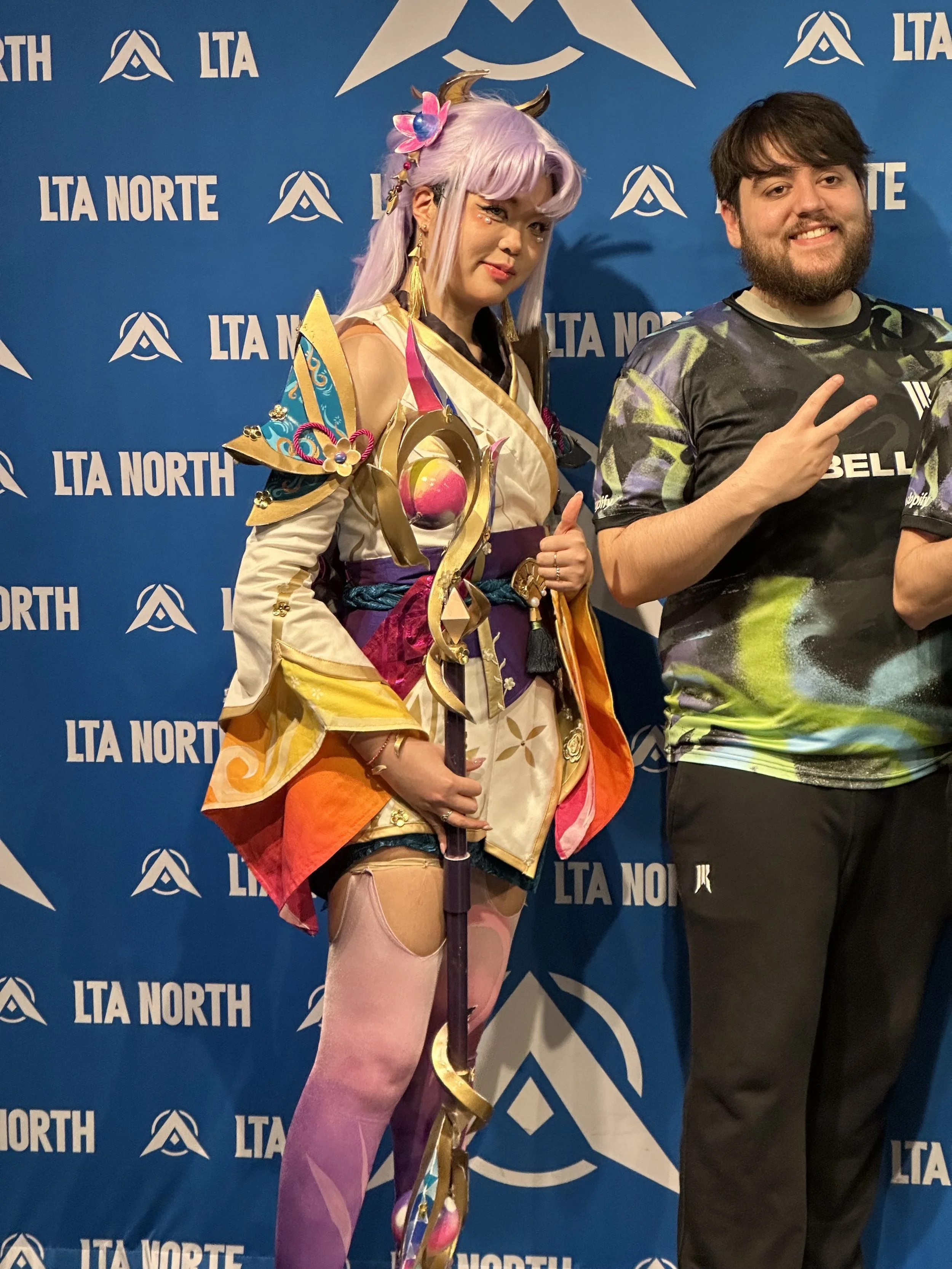 A woman in elaborate cosplay costume with pastel purple hair, gold horns, and fantasy armor holding a staff, standing next to a man making a peace sign, in front of a blue backdrop with "LTA NORTH" logo.
