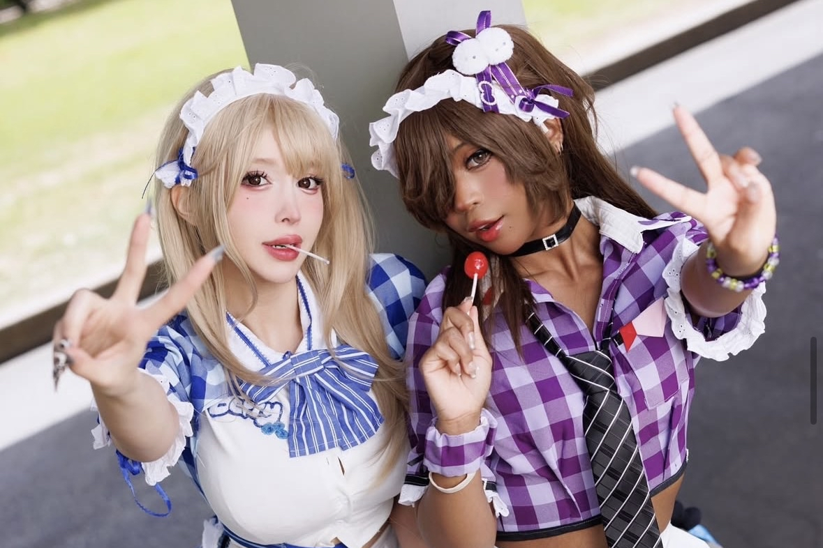 Two young women dressed in maid-inspired costumes with plaid shirts, headbands, and accessories posing and making peace signs. One is holding a red lollipop, and the other has a lollipop in her mouth.