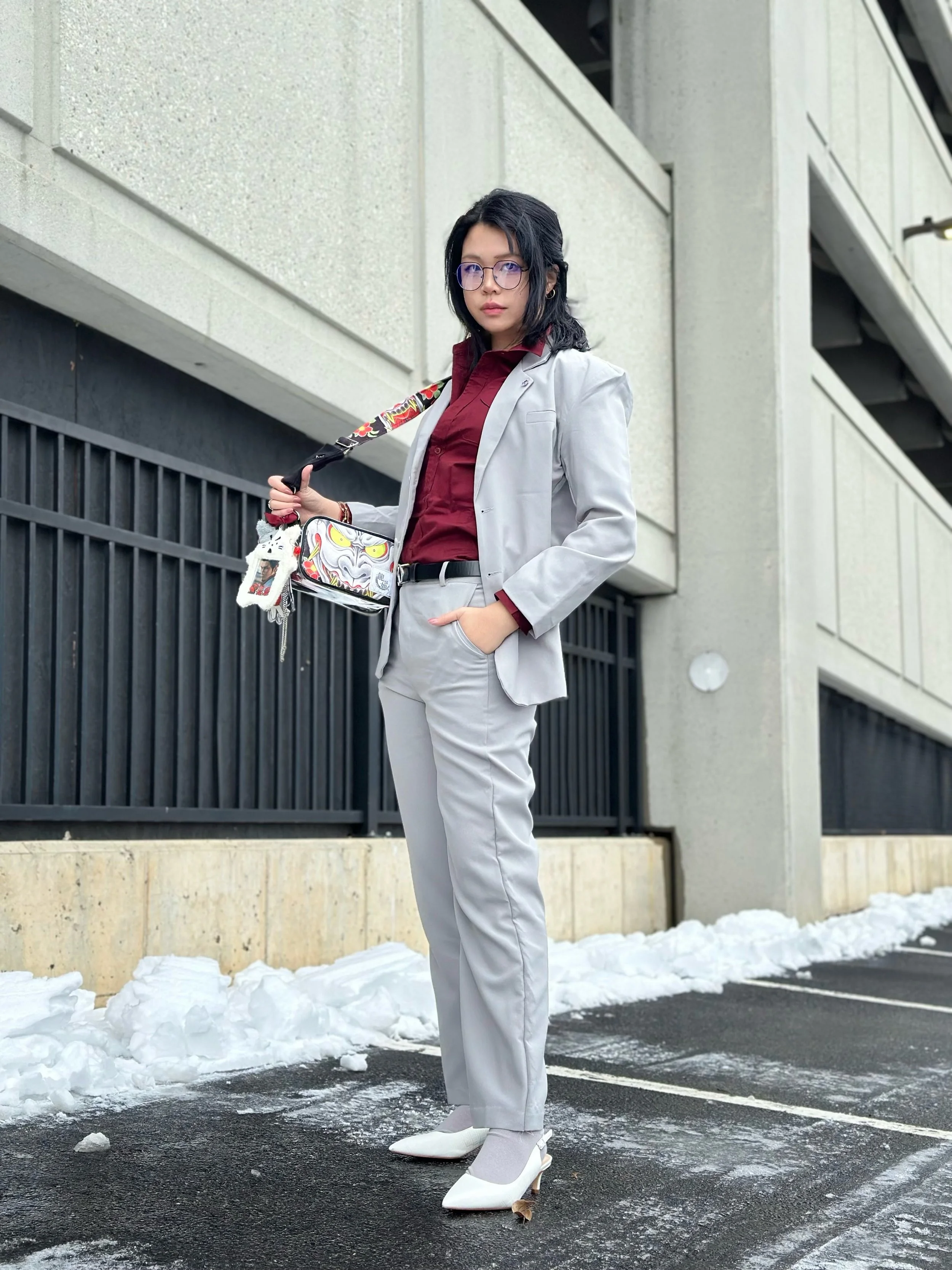 A woman in a light gray suit with a maroon shirt and white heels standing outside in a parking lot with snow, holding a colorful bag and a skateboard.