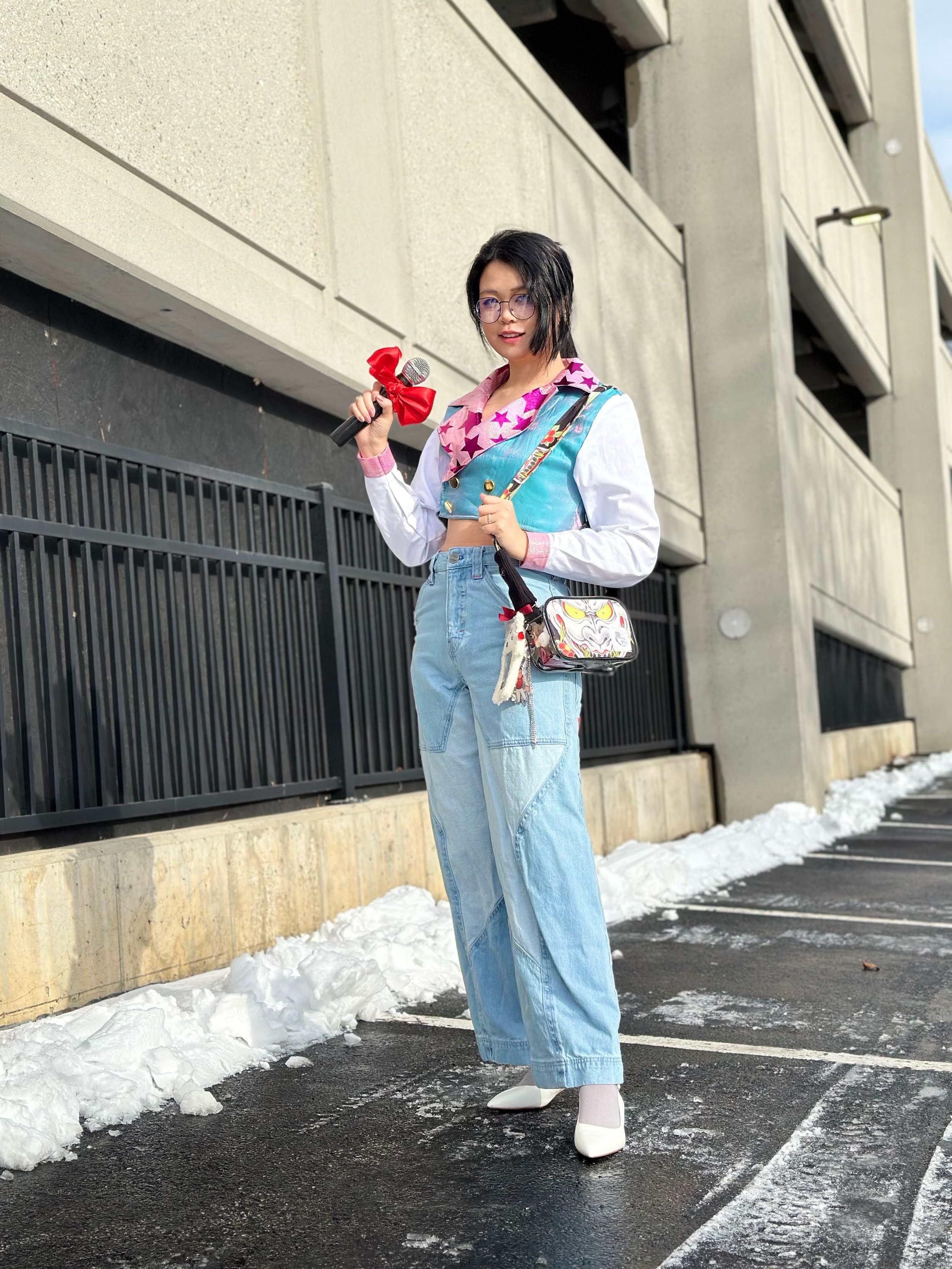 A woman standing outdoors in a parking lot with patches of snow, holding a microphone with a large red bow attached. She is wearing glasses, a colorful star-patterned scarf, a cropped blue vest, white shirt, light blue high-waisted jeans, white high 