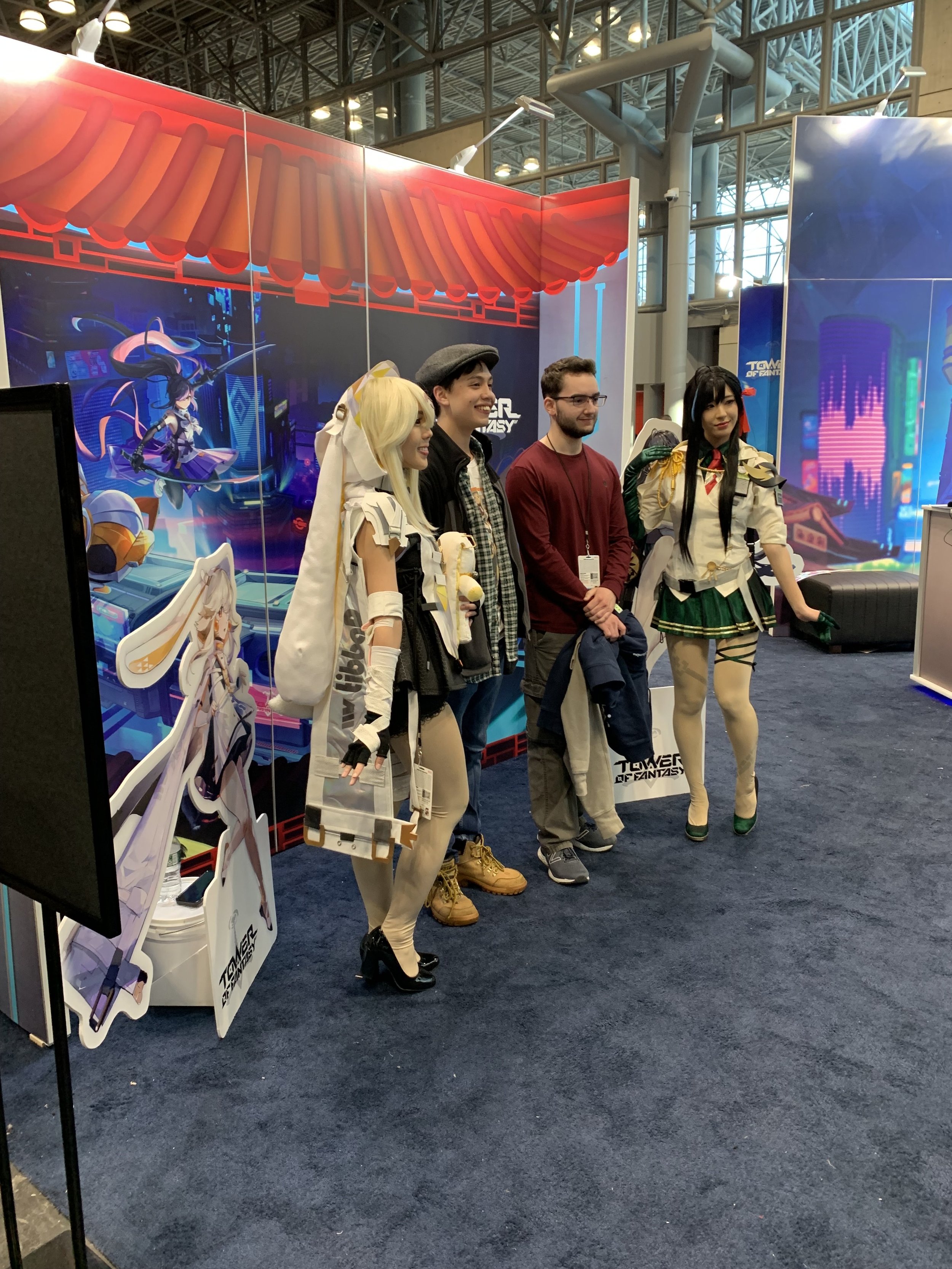 Four people standing together at a gaming convention, two women in anime-style costumes and two men in casual clothing, near a promotional booth for the game Tower of Fantasy.