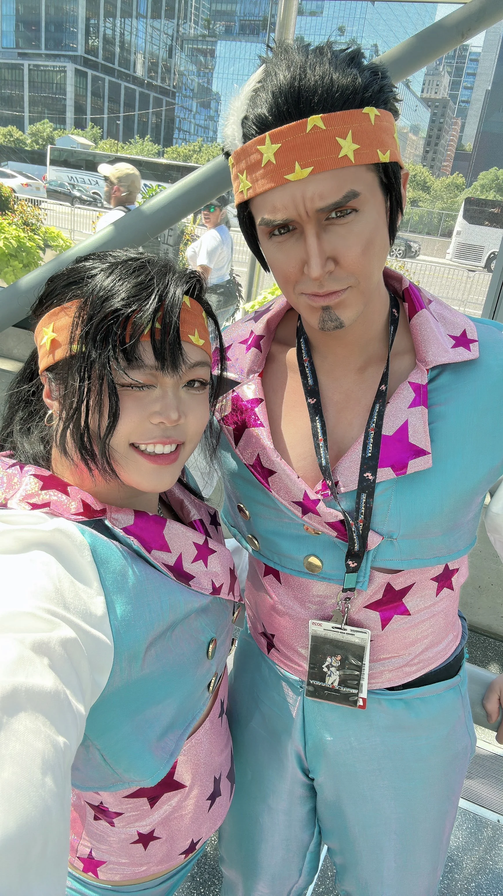 Two people dressed as Elvis Presley, one woman and one man, taking a selfie at an outdoor event with modern glass buildings and trees in the background. Both are wearing colorful outfits with star patterns, matching headbands, and lanyards with badge