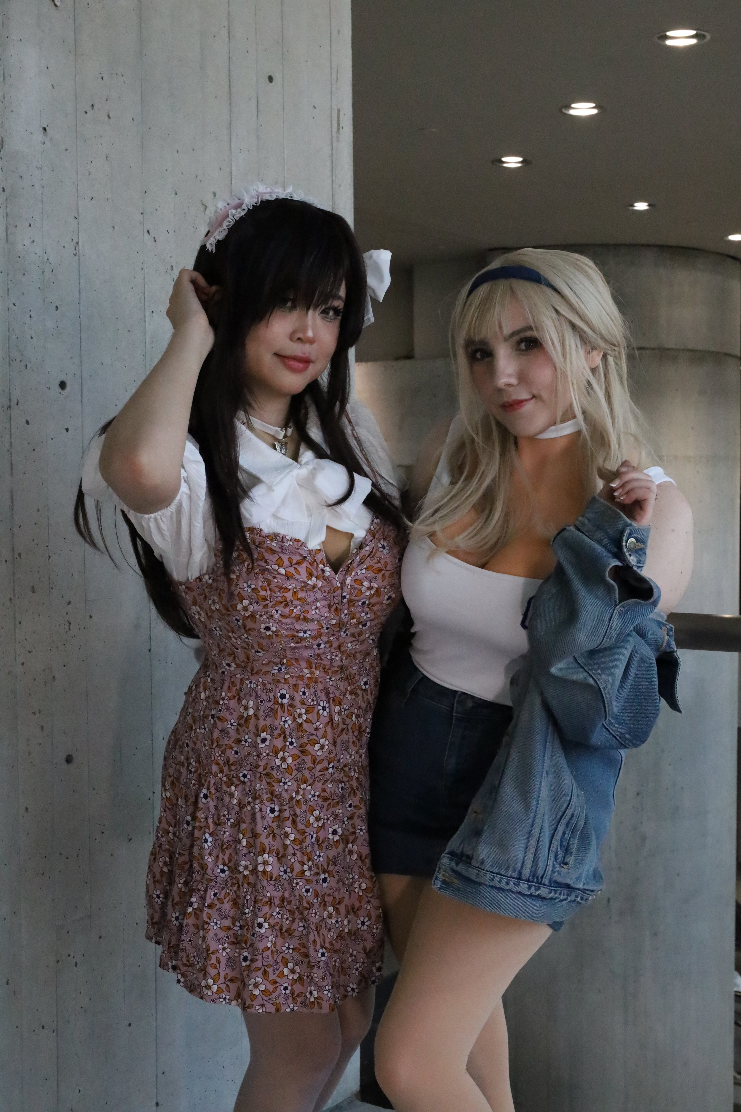 Two young women standing close together indoors, one with long dark hair in a floral dress and the other with long blonde hair in a white tank top and denim jacket.