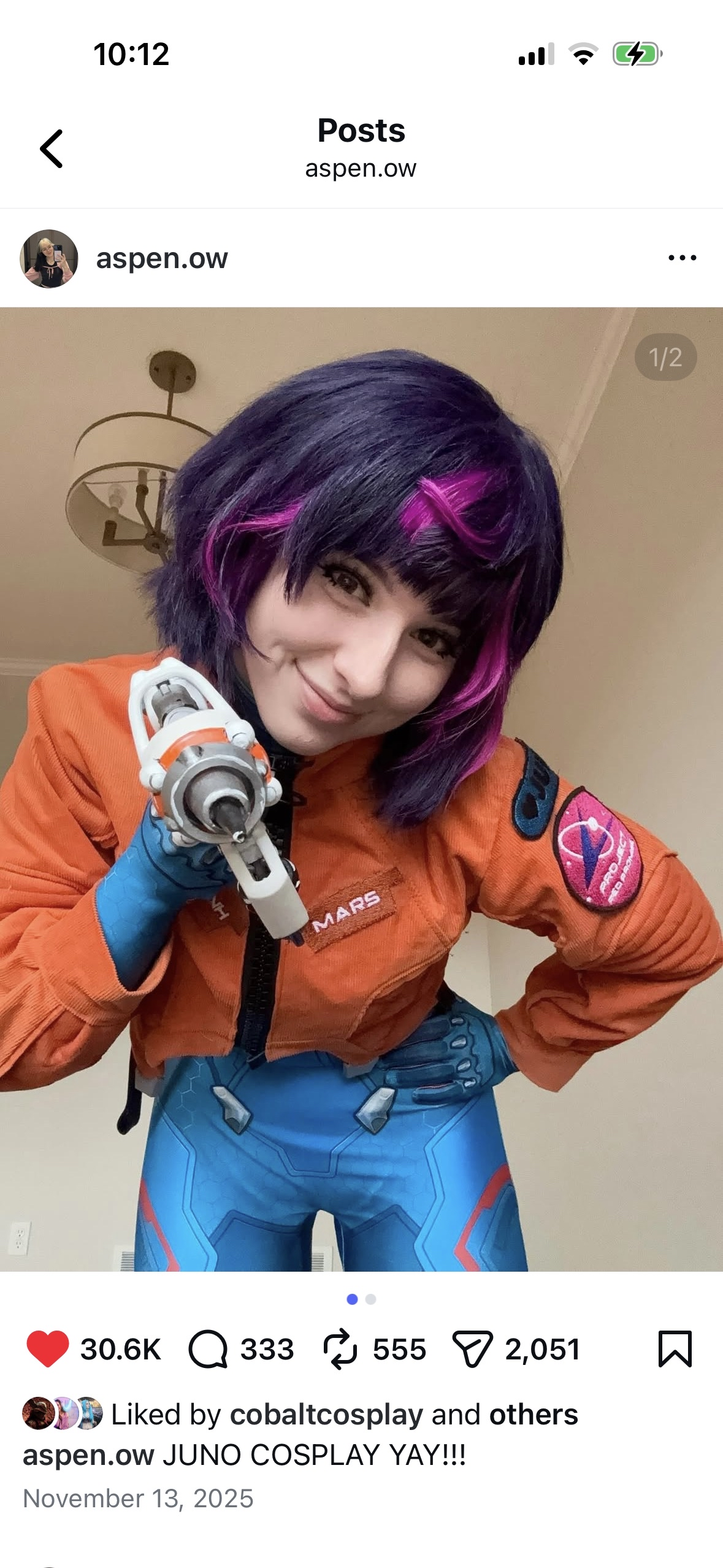 A woman in a cosplay costume inspired by the character Juno from the video game Mass Effect, wearing a space suit with patches and a futuristic gun. She has short, styled purple hair with pink streaks and is smiling.