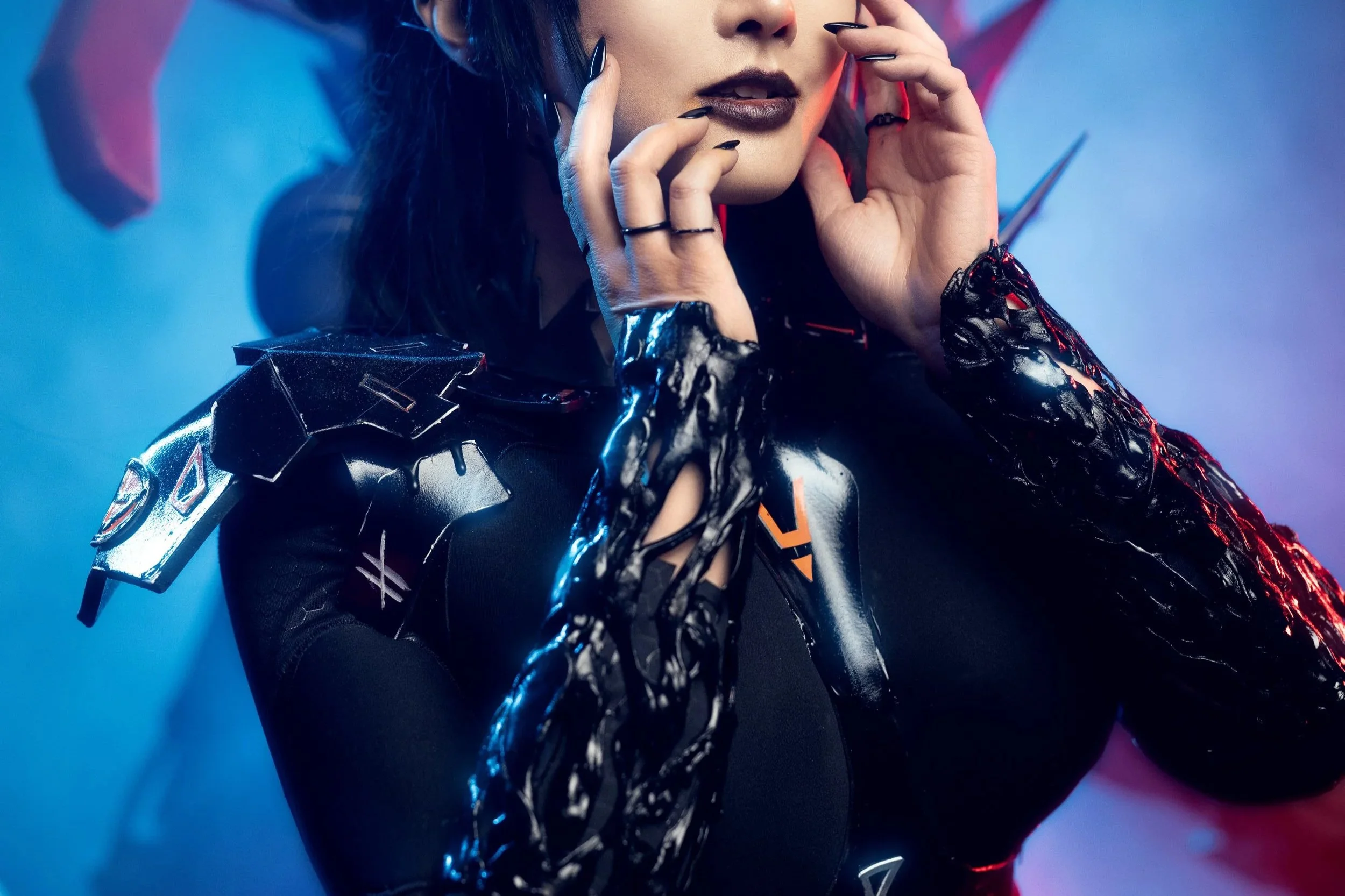 A woman with dark hair and dark lipstick, dressed in a futuristic black outfit with armor-like shoulder details, is touching her face with her hands, which have black rings and metallic gloves. The background features red and blue lighting, creating a sci-fi atmosphere.