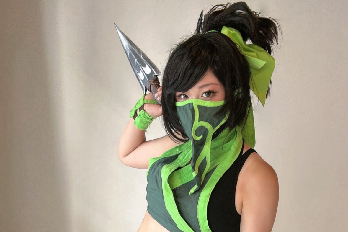 A woman dressed in a green and black ninja costume, with a matching face mask, holding a dagger, and wearing a green ribbon in her hair.